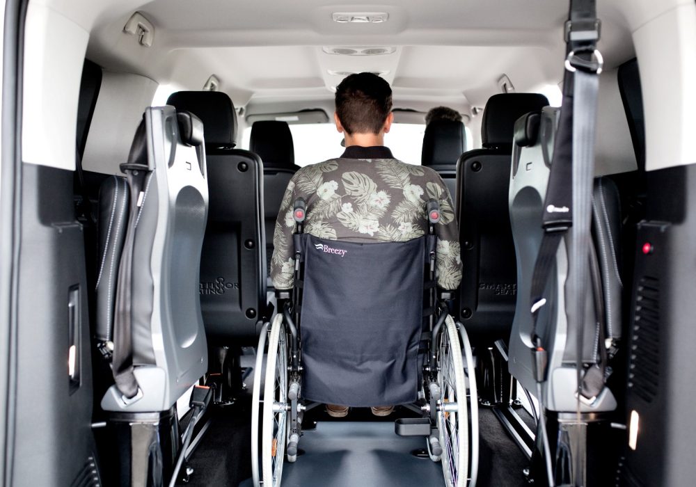 Personne en fauteuil roulant vue de dos installée à bord d'un Peugeot Traveller M TPMR au niveau de son décaissement avec sa rampe déployée - véhicule TPMR - voiture handicap